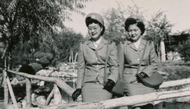 Two Nisei women in cadet nurse uniforms sitting on a wooden railing.